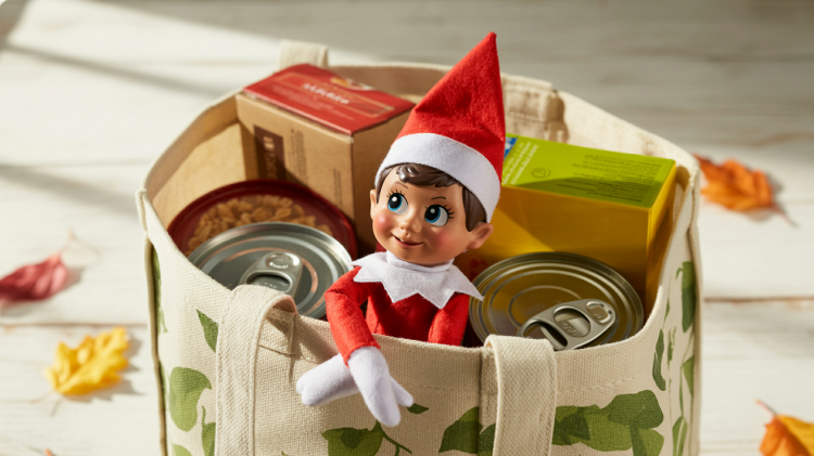 an elf on a shelf doll peeking out from inside a large, reuseable grocery bag filled with non-perishable food